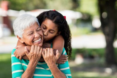 A loving teen girl embracing and kissing her happy grandmother from behind and holding each other outdoors. - Fremstad Law