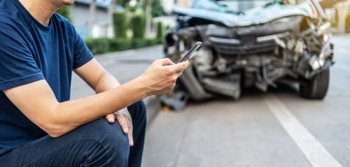 man dialing a number on his cell phone sitting in front of car accident site - in a car accident concept